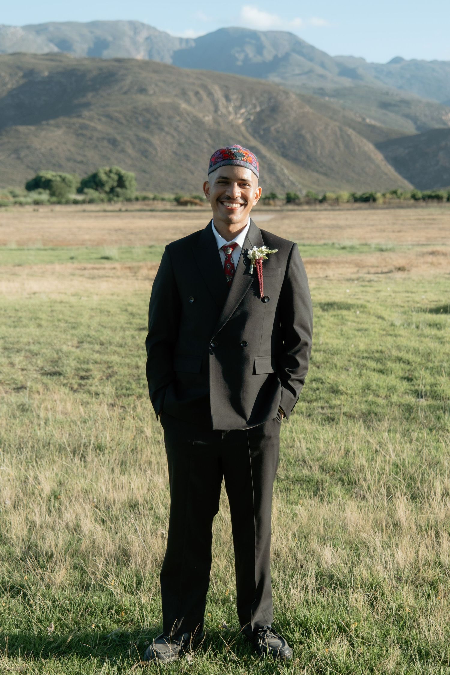 Groom in black suit and traditional red kufi cap smiles in open field with mountain backdrop.