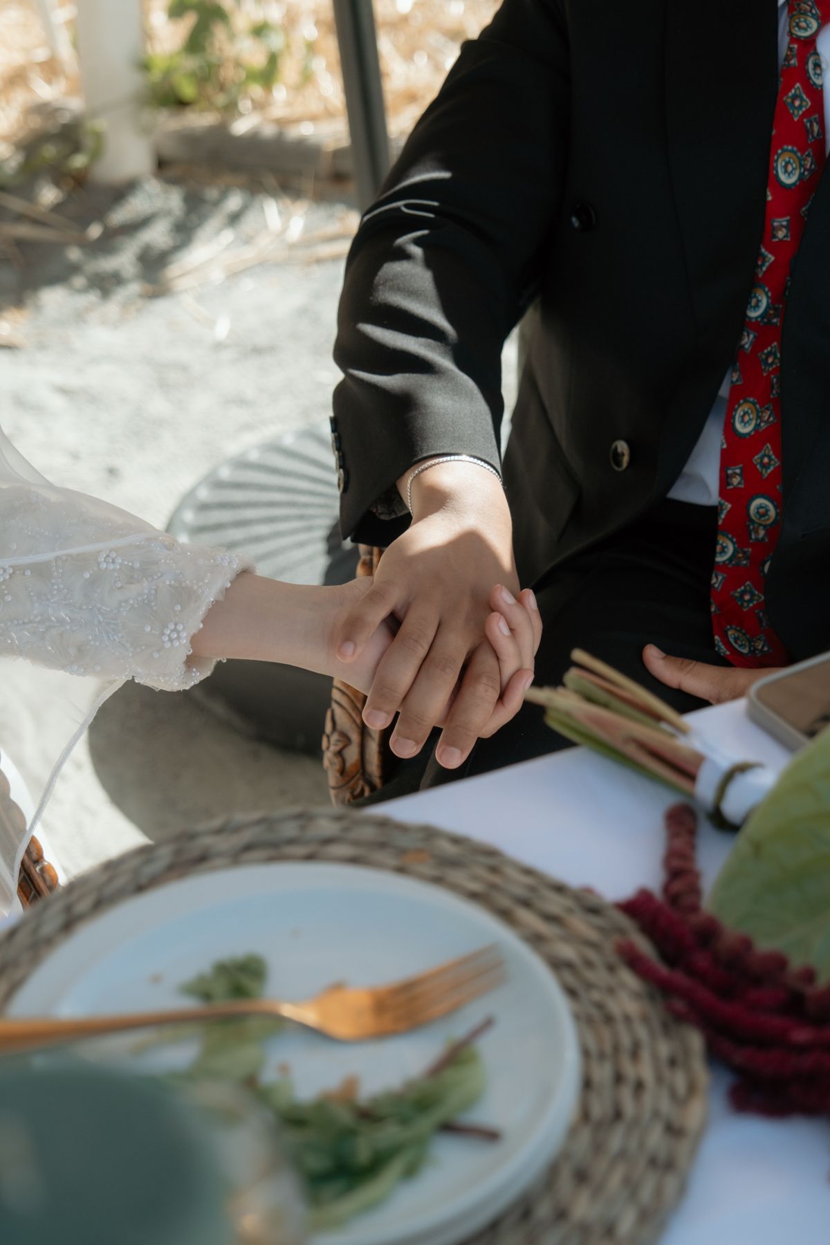 Bride and groom holding hands at wedding reception table with gold cutlery and woven placemats.