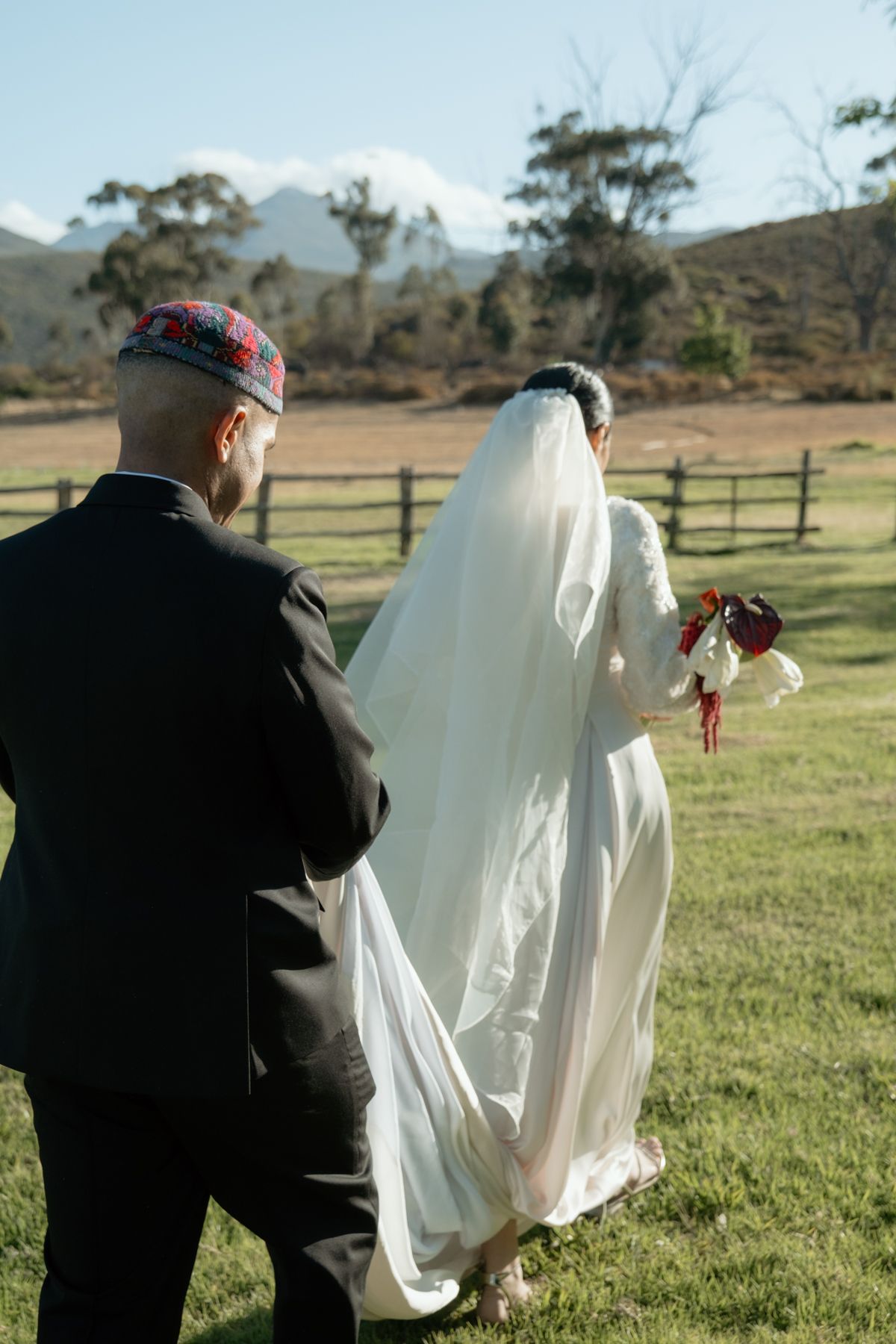 Muslim bride in white gown and veil walks with groom in black suit and kufi cap across scenic field.