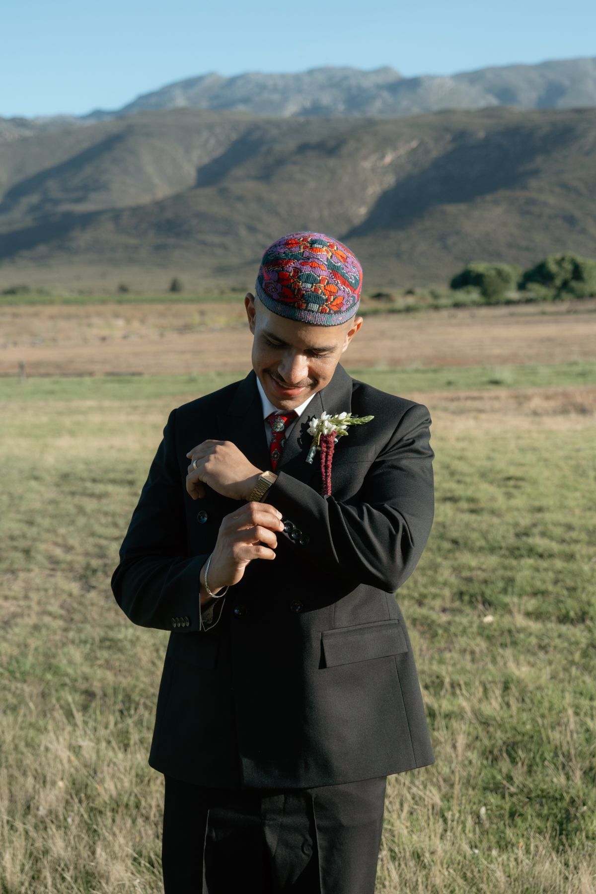 Groom in black suit and colorful kufi cap adjusts cufflinks in open field with mountain backdrop.