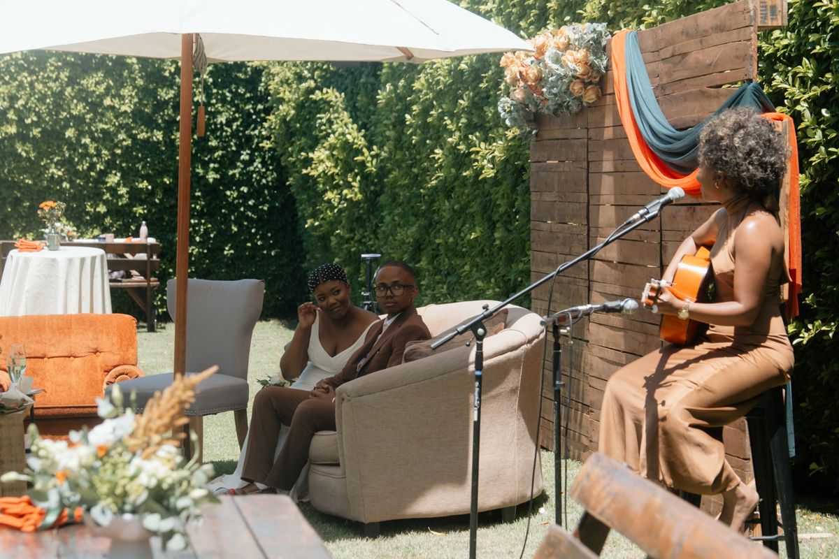 Woman playing guitar at outdoor gathering while two guests relax on sofa in sunny backyard setting.