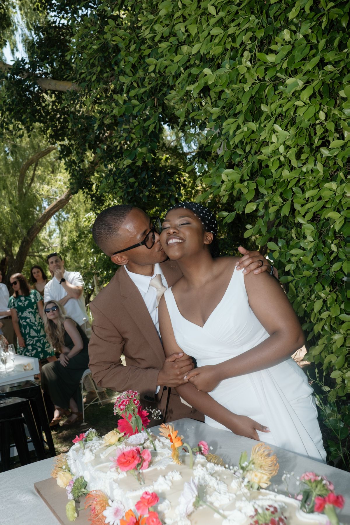 Bride and groom share a kiss while cutting wedding cake at outdoor garden reception.