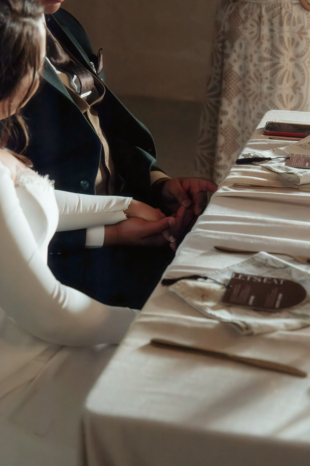 Bride and groom hold hands tenderly at decorated wedding reception table in warmly lit venue.