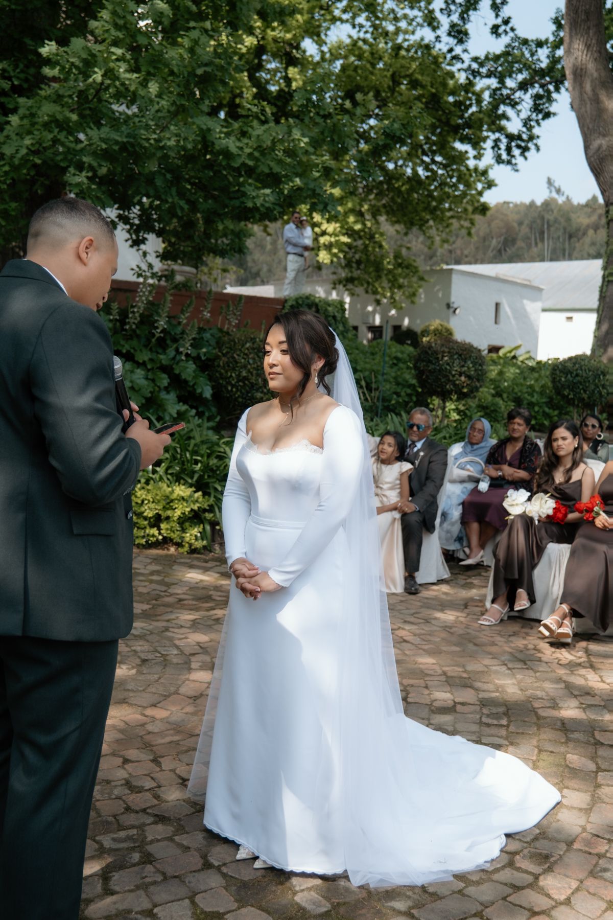 A bride in a long white gown and veil stands at an outdoor wedding ceremony exchanging vows with the groom.