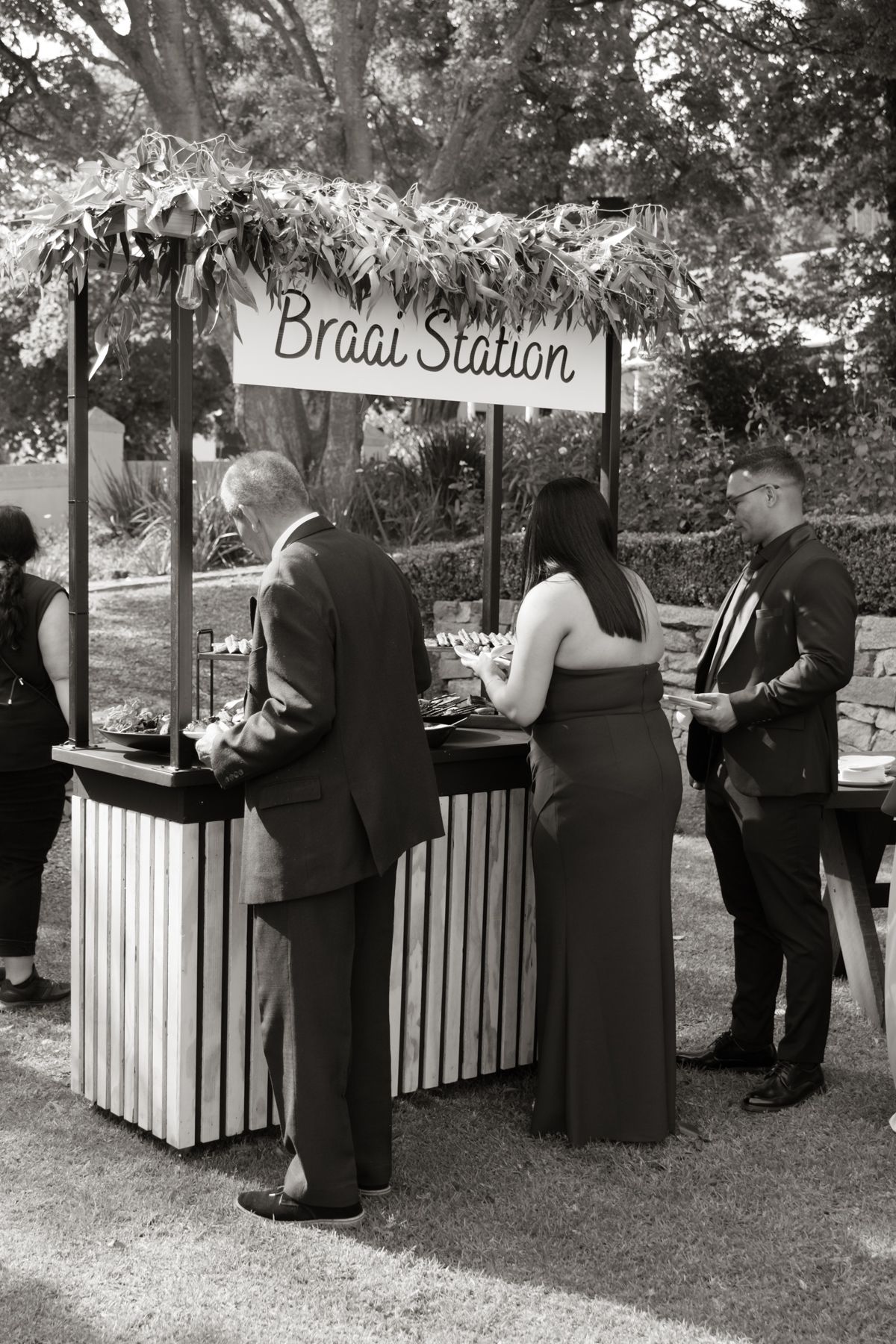 Wedding guests browse outdoor Braai Station food stand decorated with greenery at garden reception.