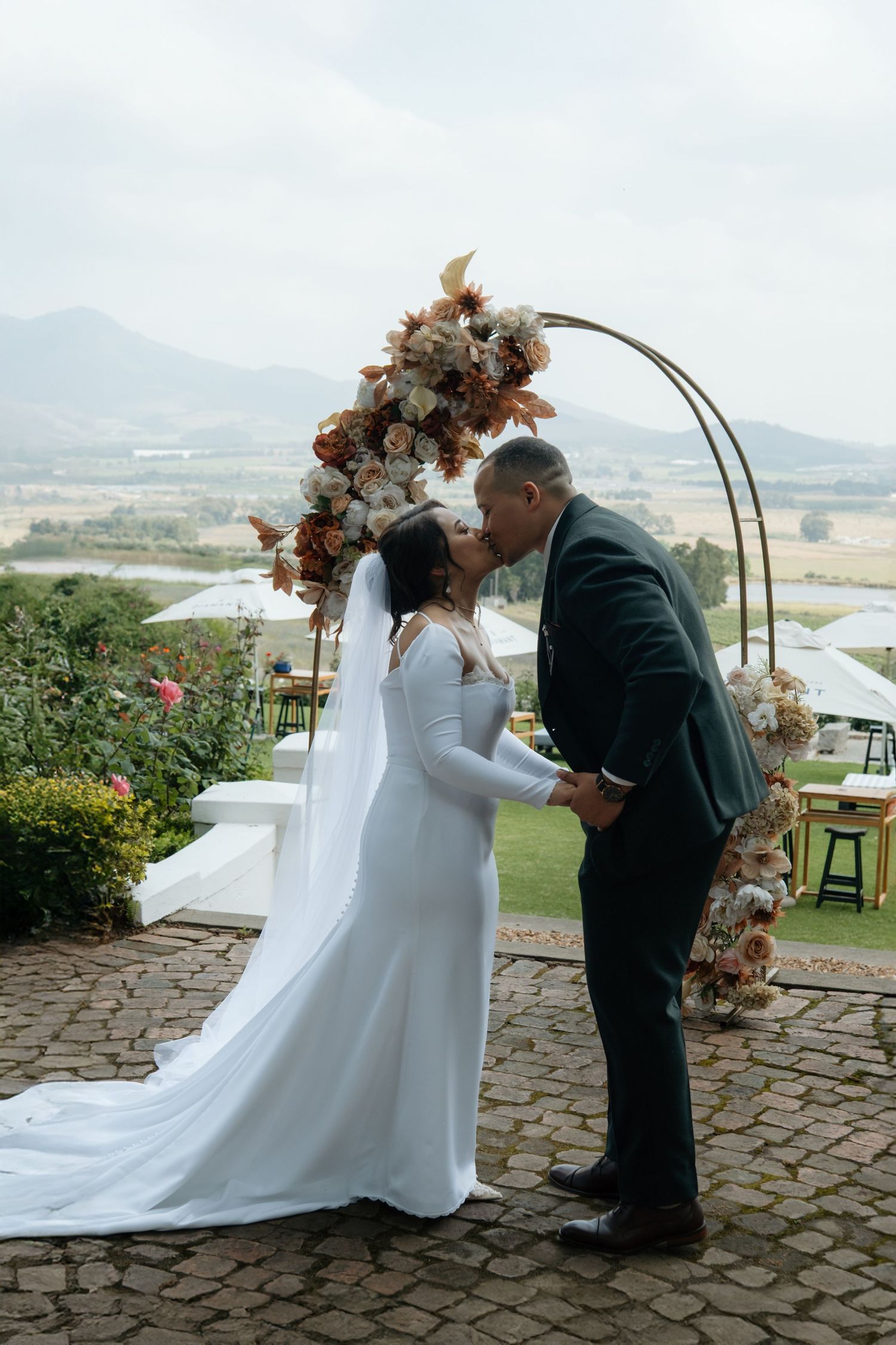 Bride and groom share first kiss under floral arch with mountain backdrop at outdoor wedding ceremony.