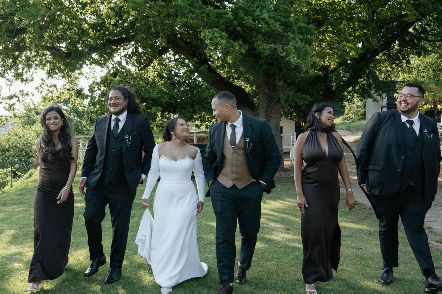 Bride and groom walking with smiling wedding party dressed in dark attire on lush green lawn.