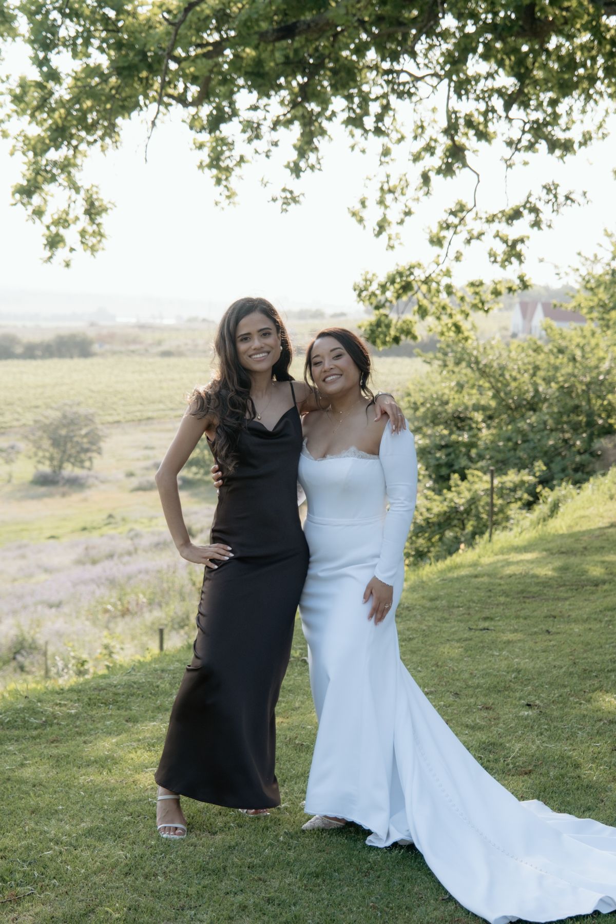 Bride in white long-sleeve gown posing with bridesmaid in black satin dress under green tree with scenic backdrop.