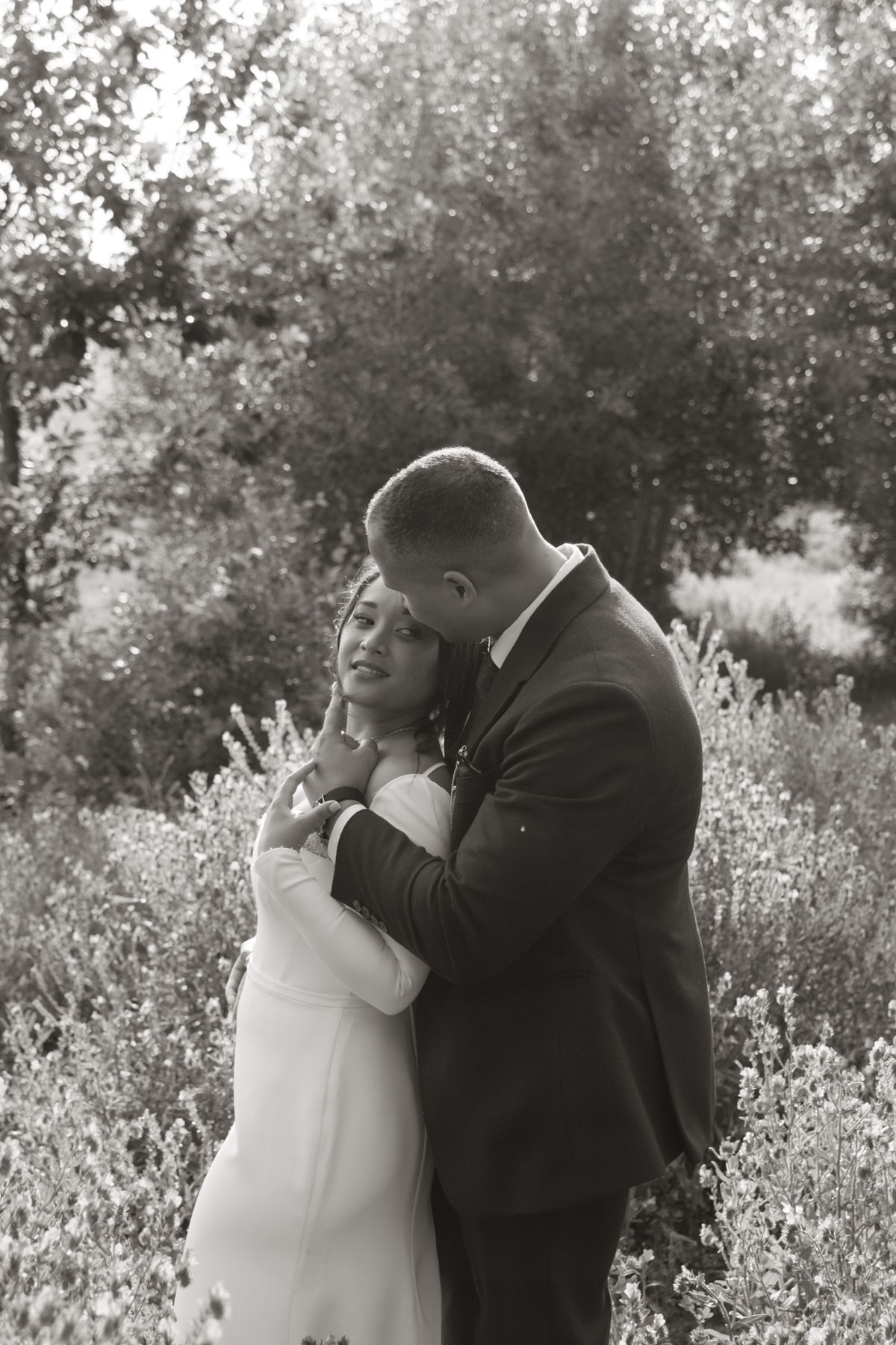 A groom embraces his bride from behind in a black and white portrait among lush greenery.