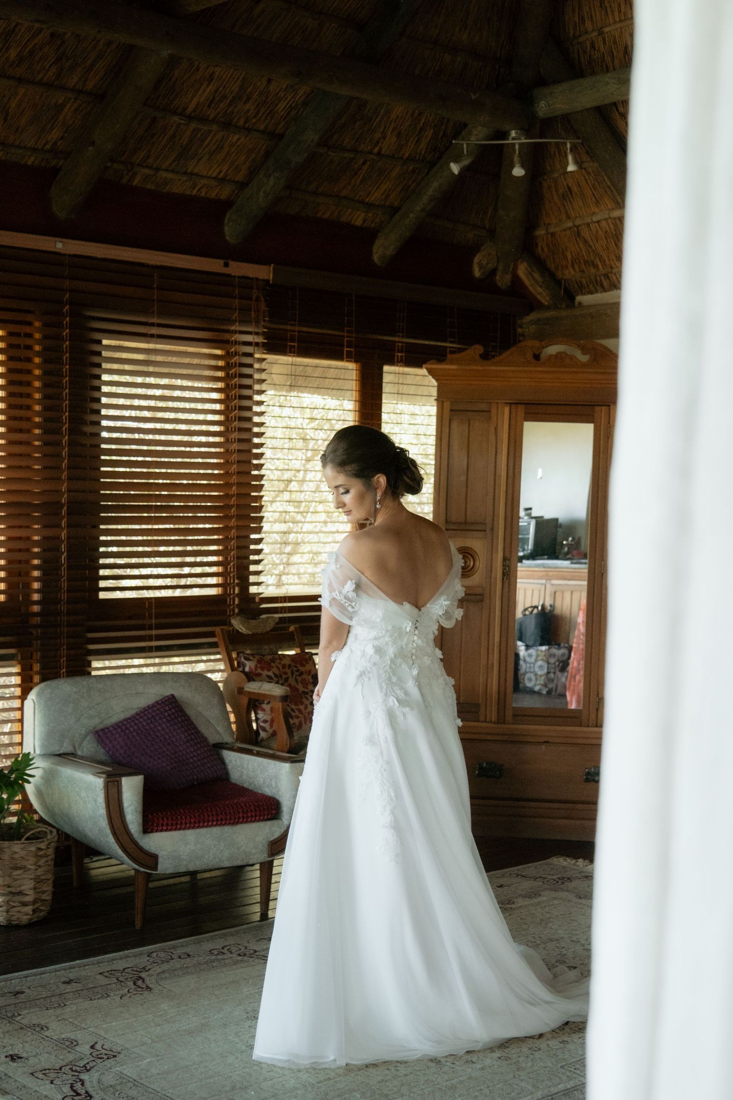 Elegant bride in off-shoulder white lace wedding gown posing in a rustic thatched roof room.