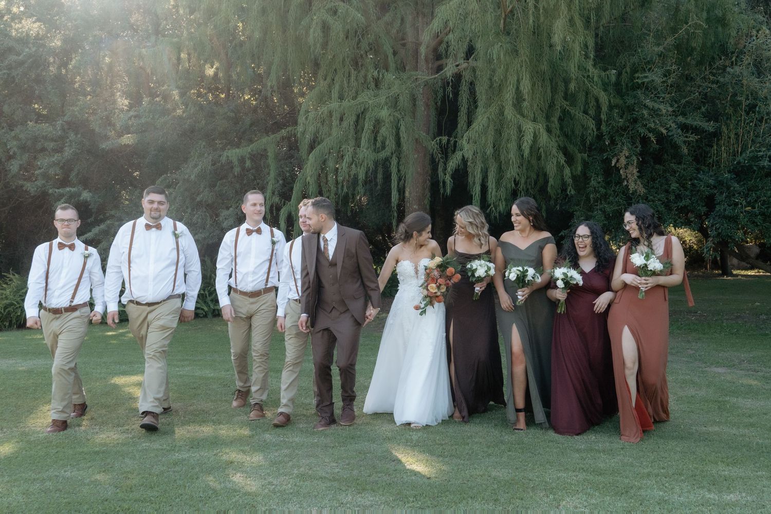 Wedding party walks together outdoors, bridesmaids in burgundy dresses and groomsmen in suspenders.