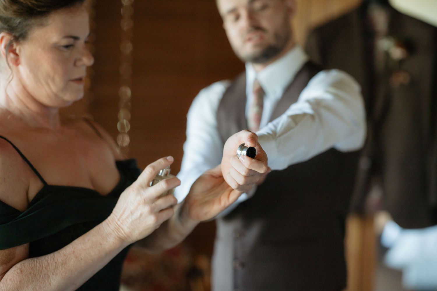 Woman in black dress fastening cufflinks on groom wearing brown vest before wedding ceremony.