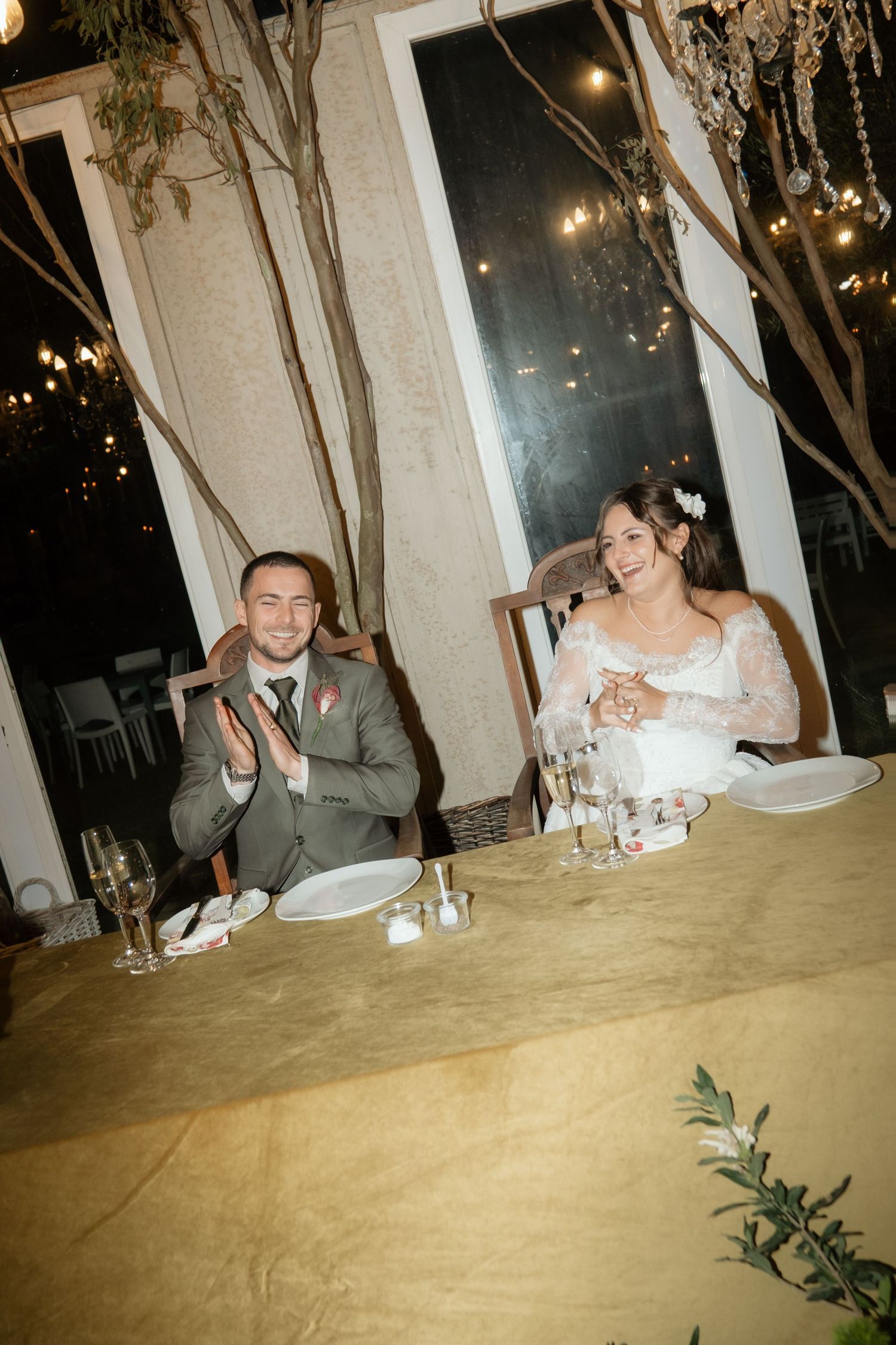 Bride and groom smiling and clapping at elegantly decorated wedding reception dinner table.