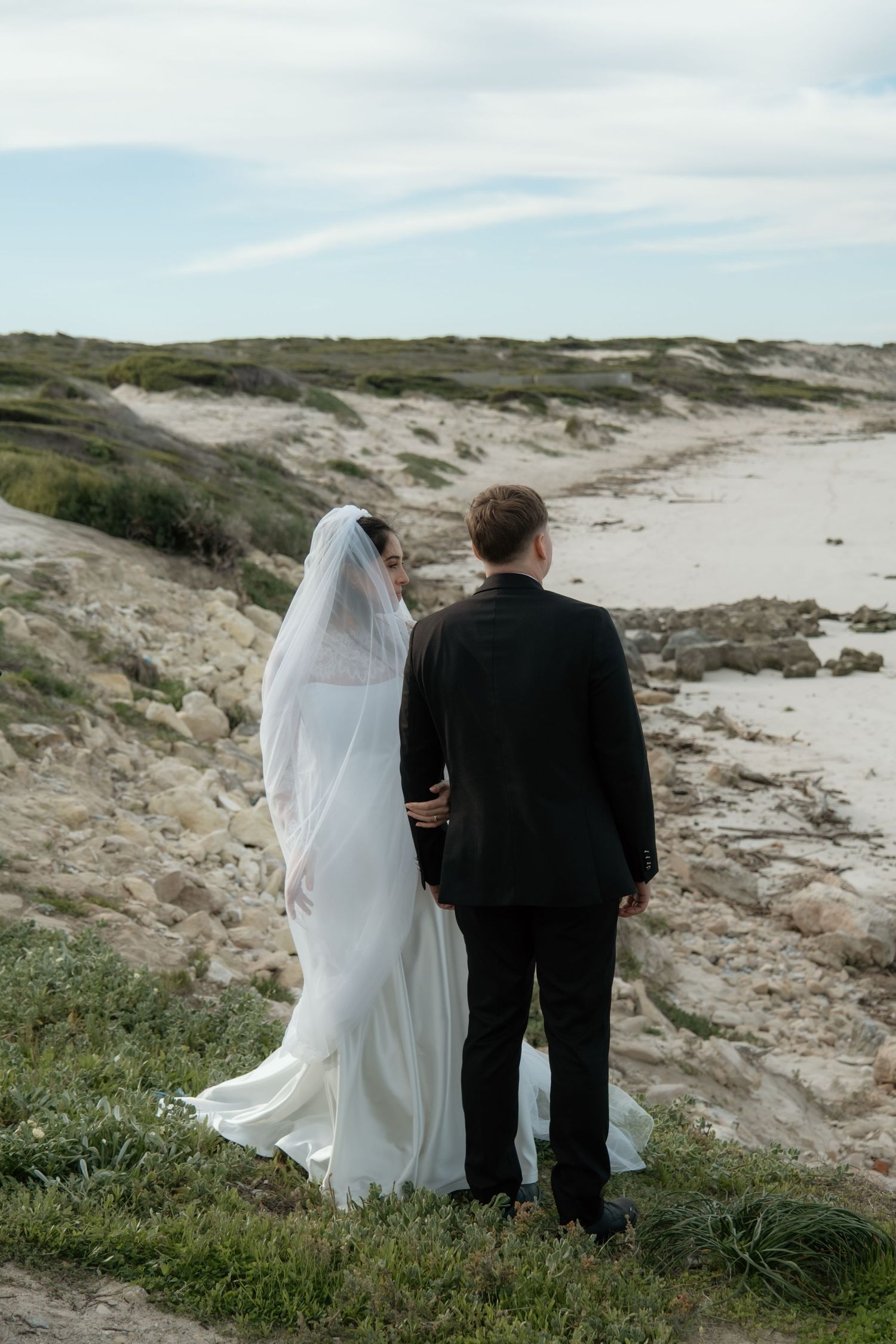 Bride and groom standing together overlooking sandy beach landscape on their wedding day.
