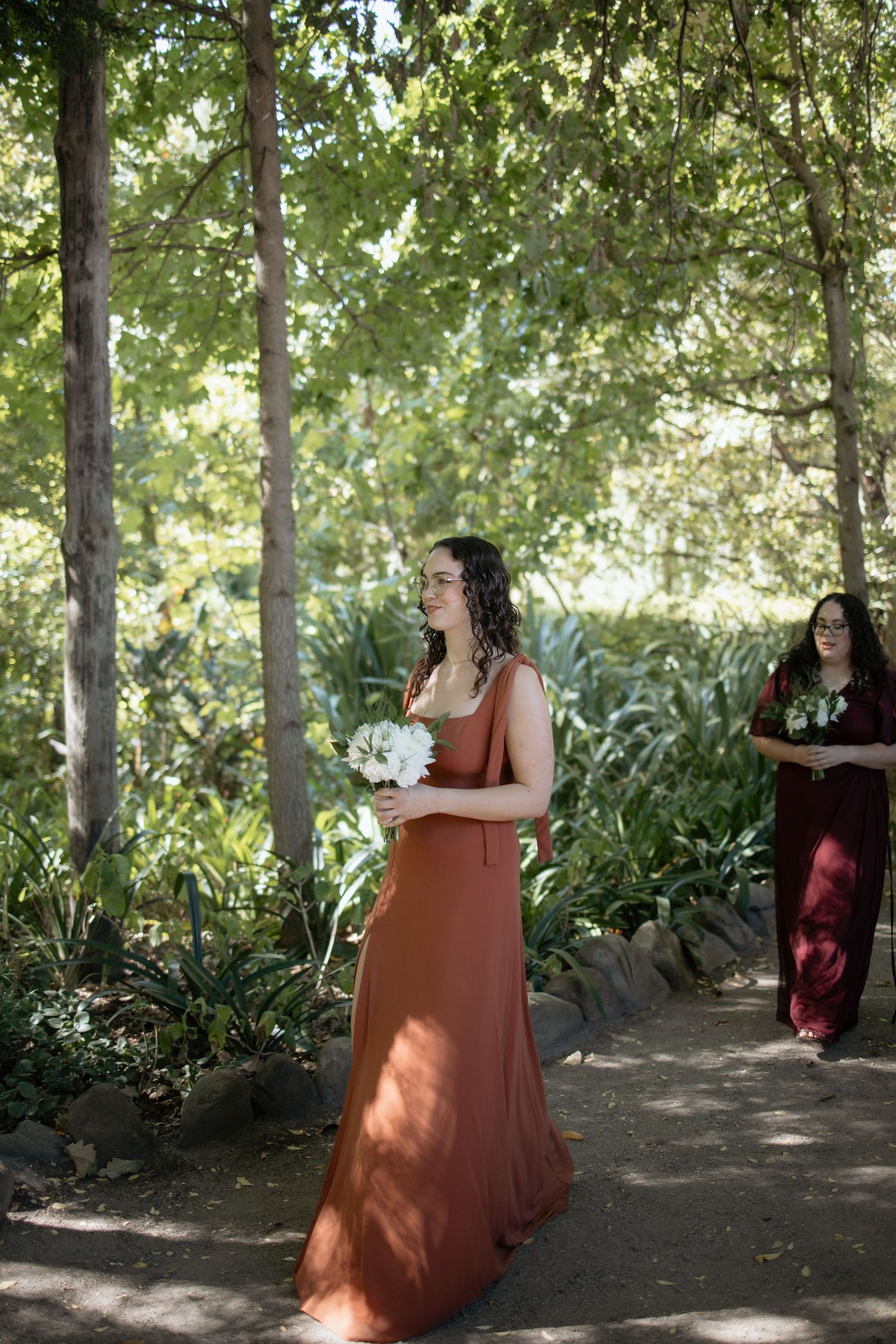 Bridesmaid in rust orange gown holds white bouquet while walking through a sunlit garden ceremony aisle.