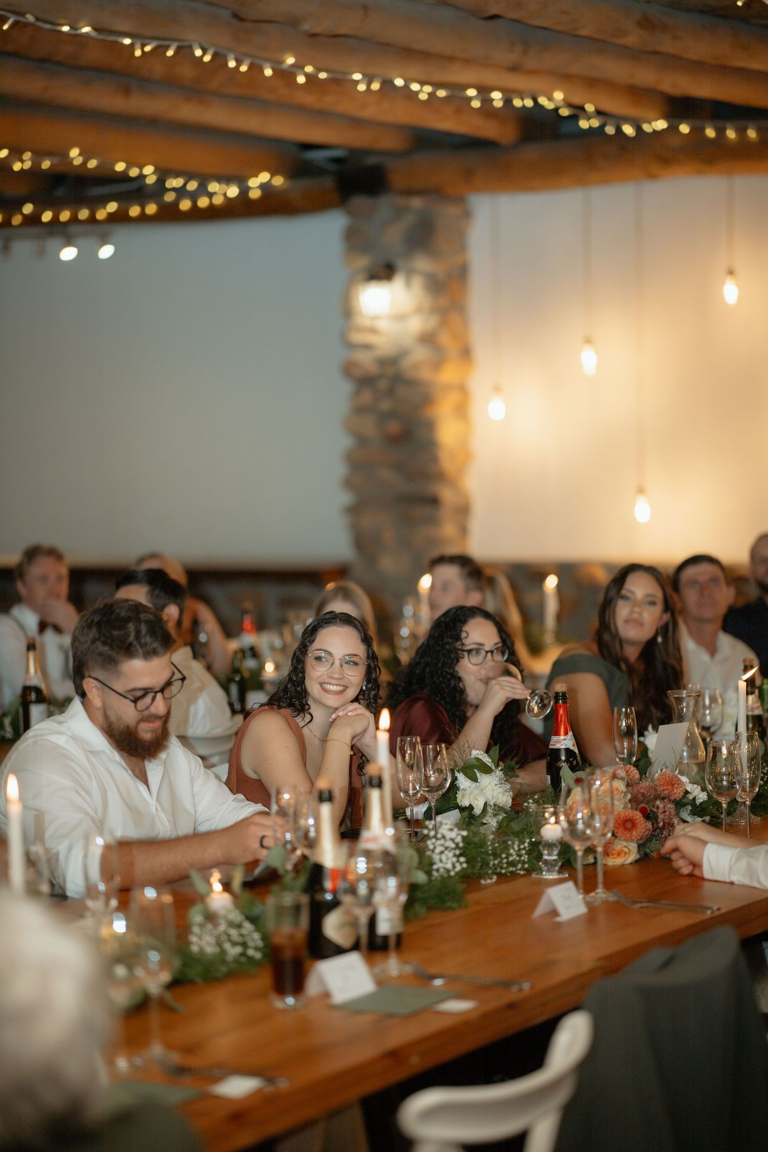 Wedding reception guests laughing at long wooden table decorated with candles and flowers under warm string lights.