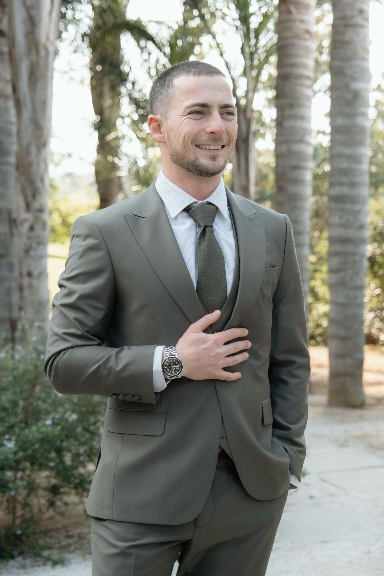 Smiling groom in olive green suit and tie poses outdoors among palm trees on his wedding day.
