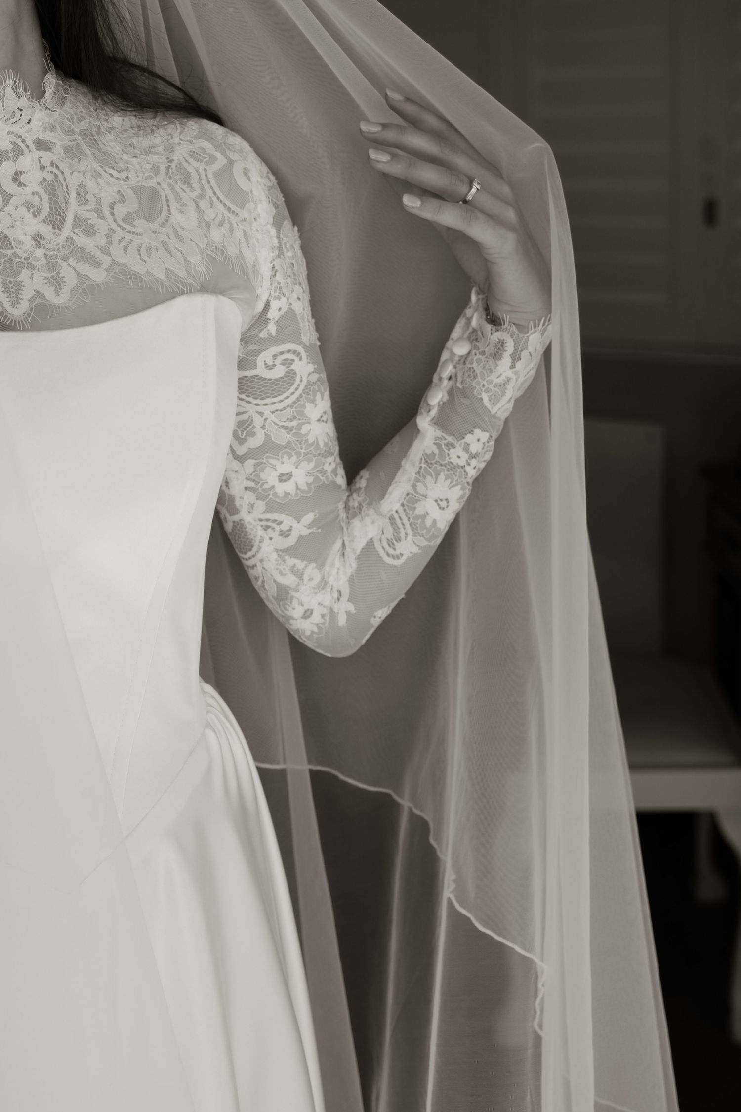 Black and white close-up of bride in lace long-sleeve wedding dress holding sheer veil.