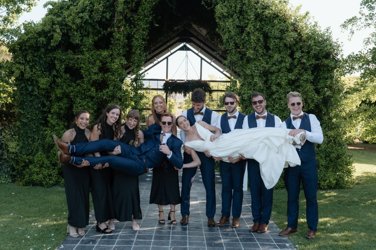 Wedding party poses playfully with groomsmen lifting bride in white dress outside garden pavilion.