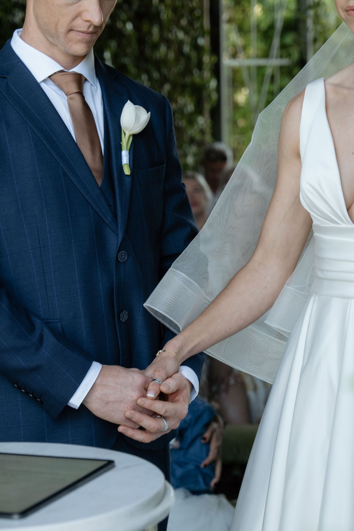 Bride and groom exchange rings during wedding ceremony, groom in navy suit with white boutonniere.
