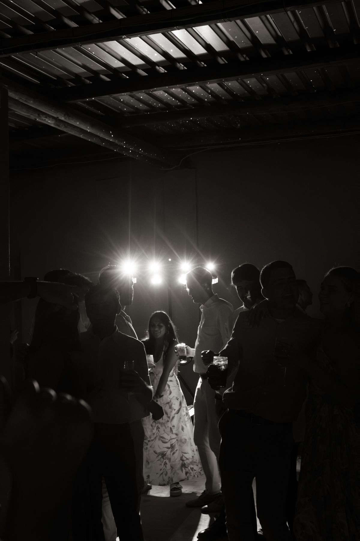 Black and white photo of wedding guests dancing under bright stage lights in a darkened reception venue.