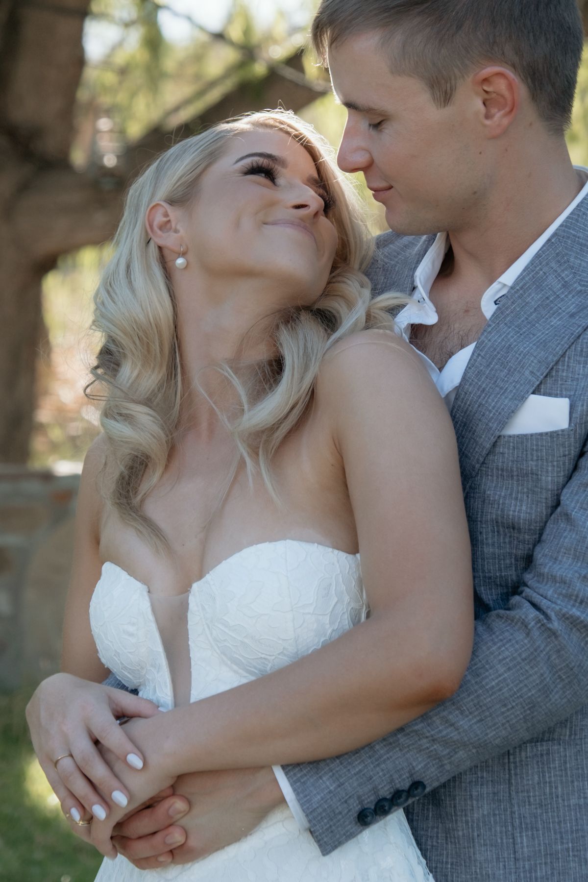 Blonde bride in strapless white gown gazes lovingly at groom in grey suit outdoors under trees.