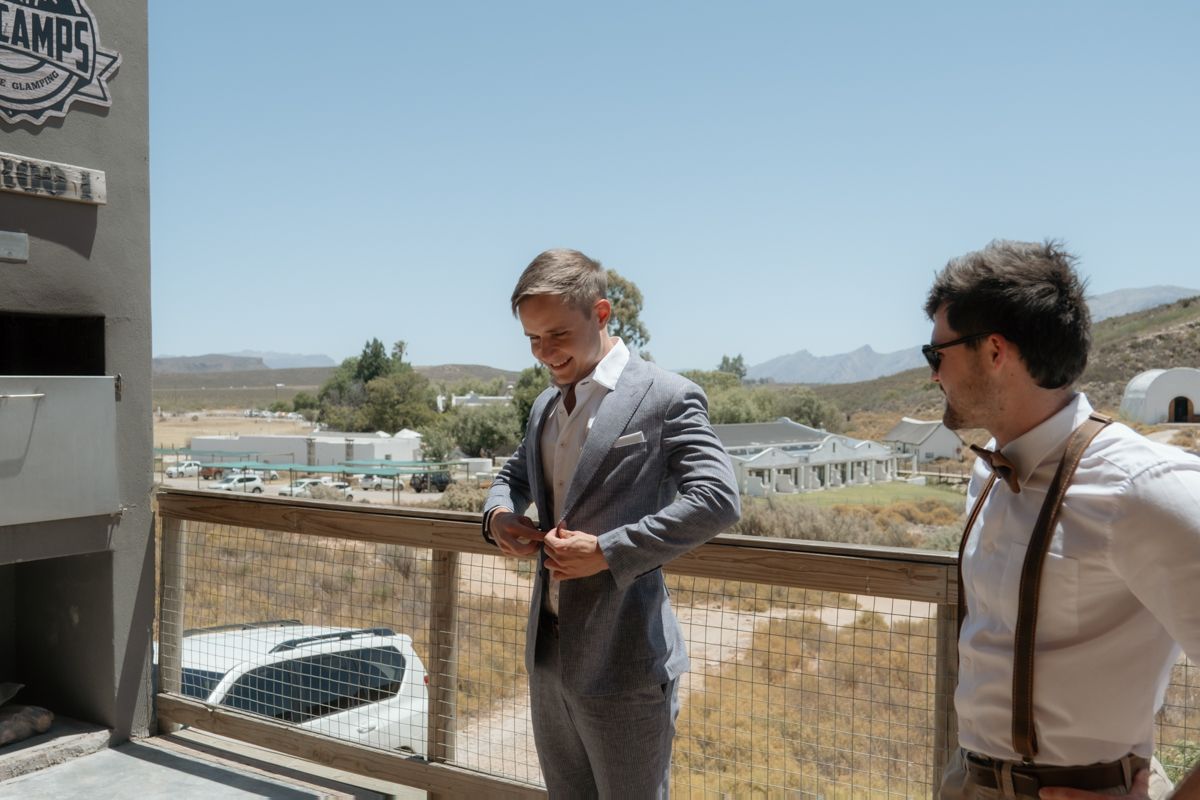 Two well-dressed men in suits stand on an outdoor balcony with a scenic desert landscape in the background.
