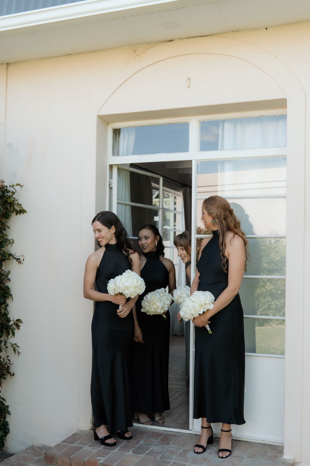 Four bridesmaids in elegant black halter dresses hold white bouquets while gathered at an arched doorway outdoors.