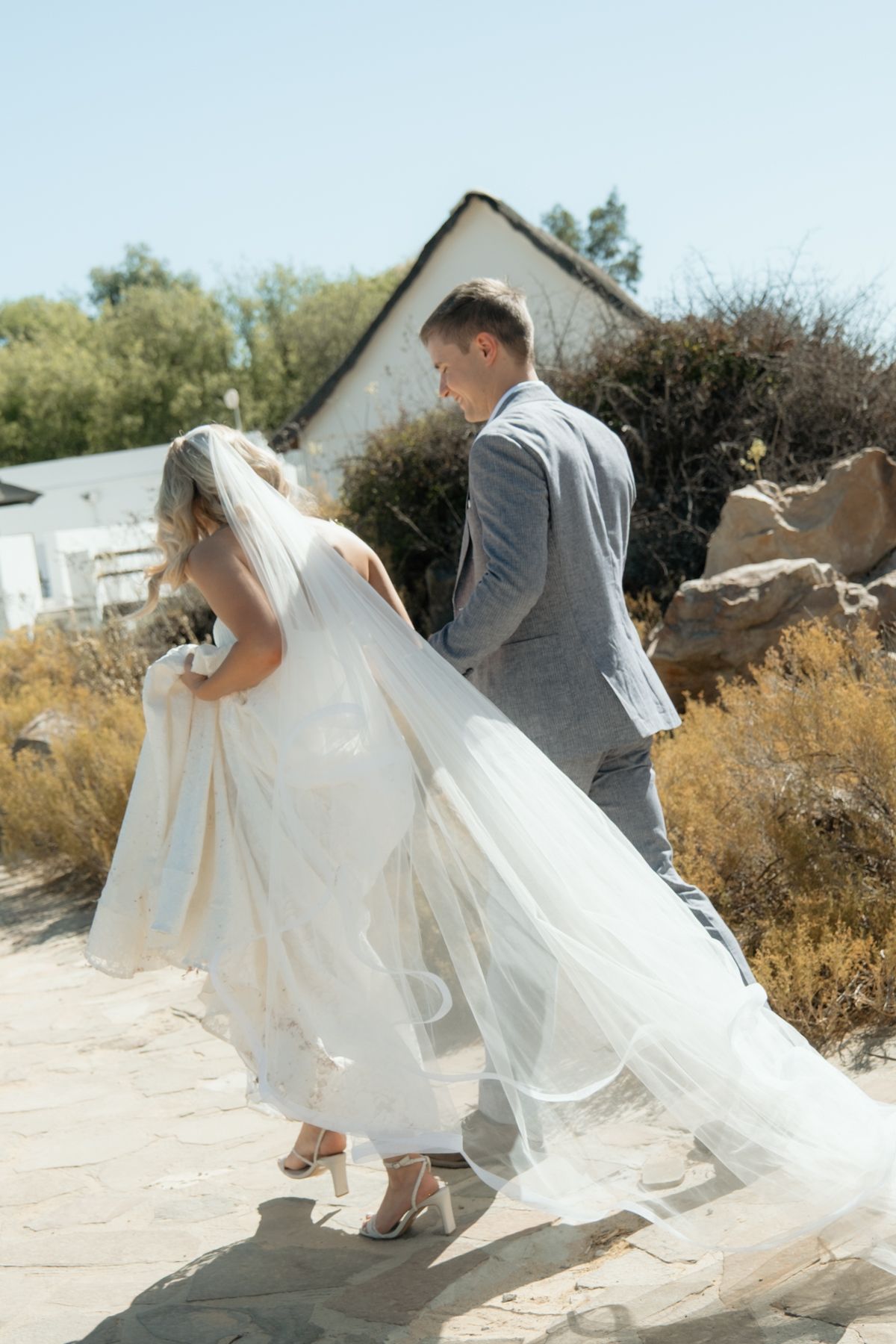 Bride and groom walking together outdoors, groom holding bride's flowing white veil on sunny day.