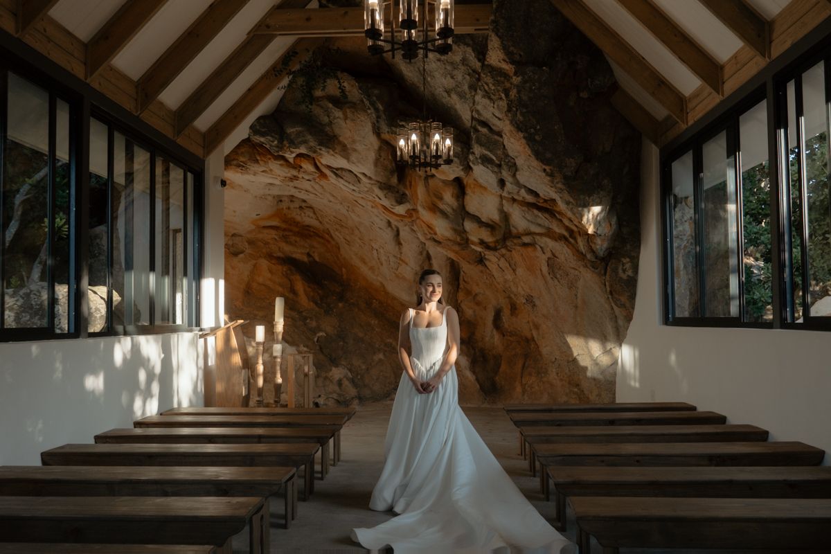Bride in a flowing white gown standing in a rustic chapel with a dramatic natural rock wall backdrop.