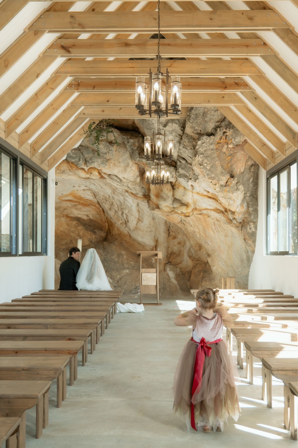 Rustic chapel interior with wooden pews, exposed beams, rock wall backdrop, chandelier, and flower girl in tutu.