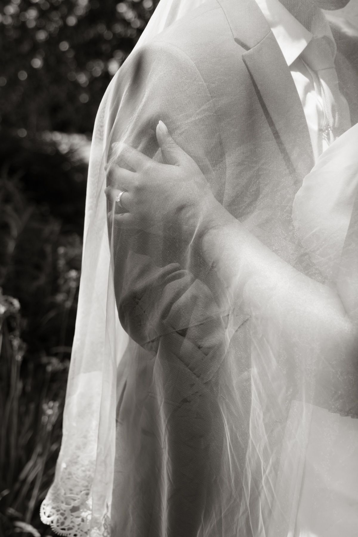 Black and white close-up of bride's hand resting on groom's arm beneath a sheer wedding veil.
