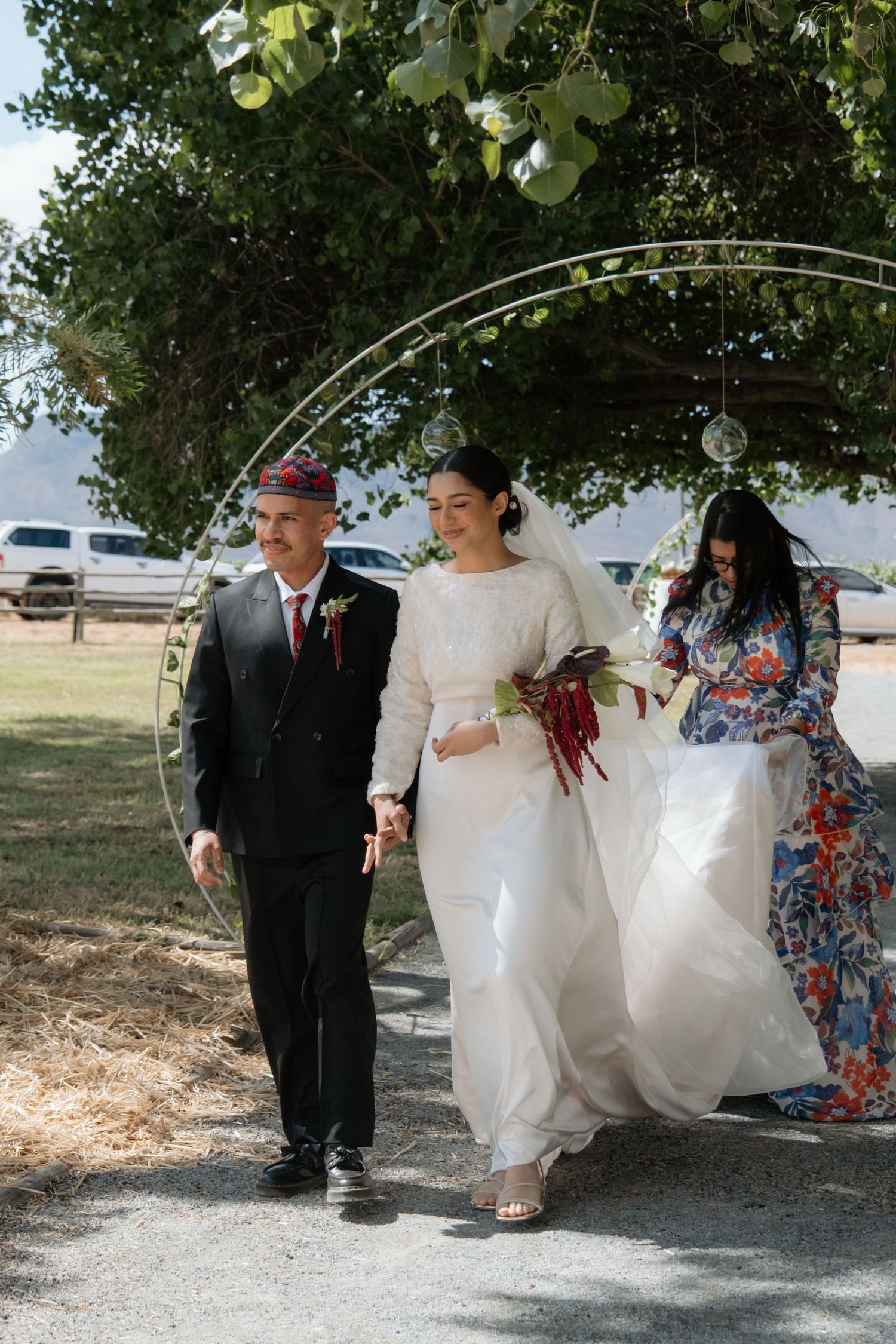 Bride in white gown walks through floral arch with father in black suit at outdoor wedding ceremony.