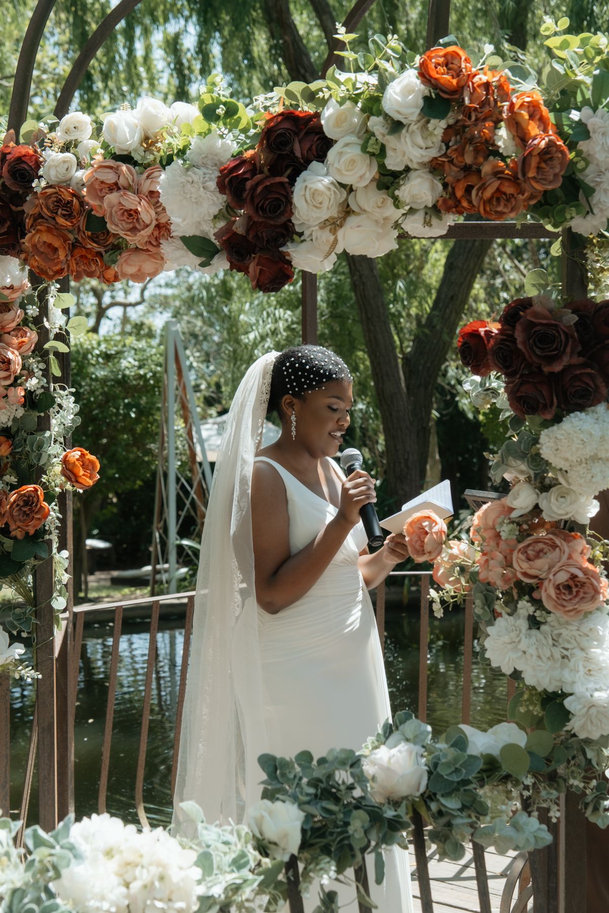 Beautiful Black bride in white gown reads vows under a floral arch of orange, red, and white roses.