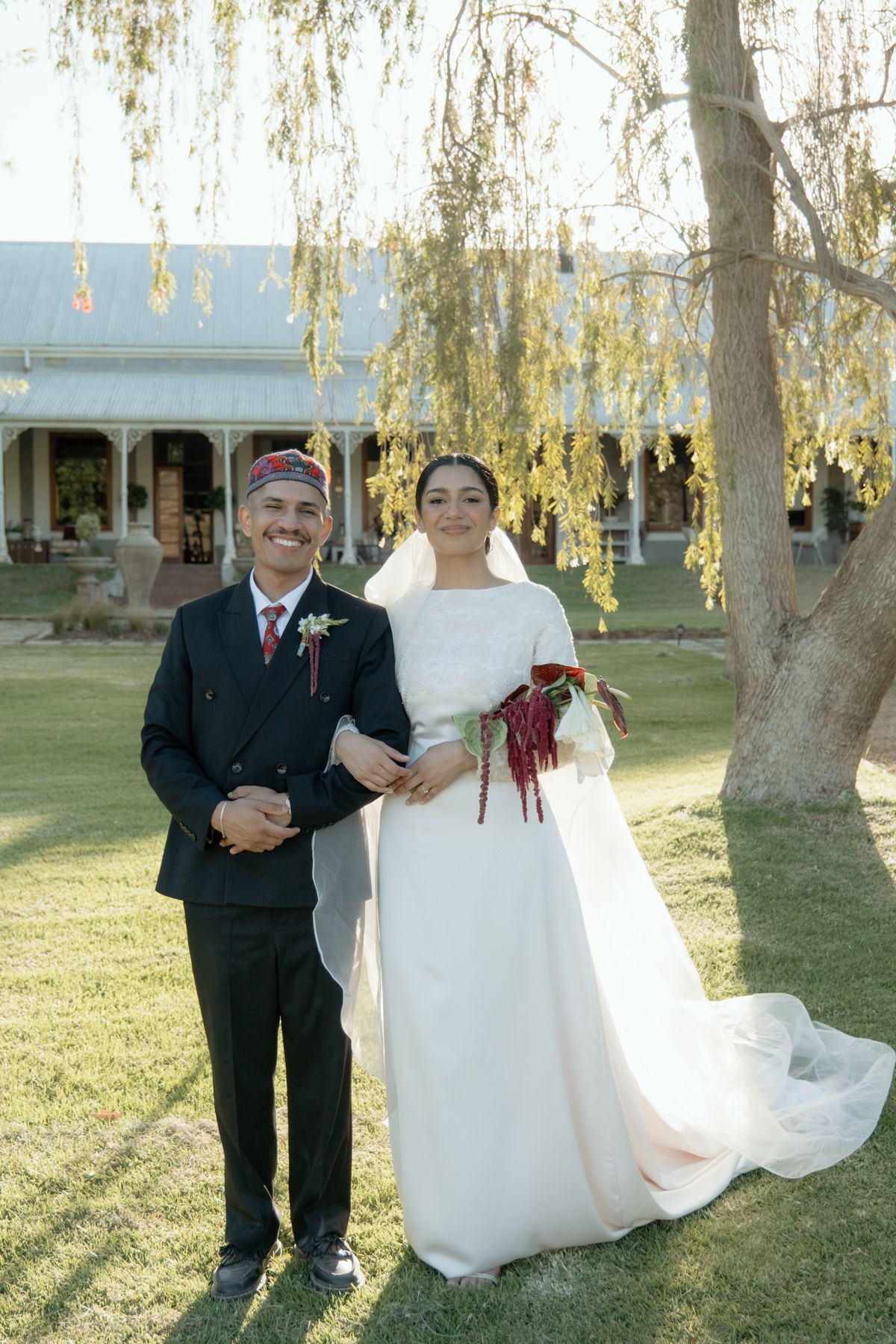 Bride and groom posing together on lawn with weeping willow tree and historic venue in background.
