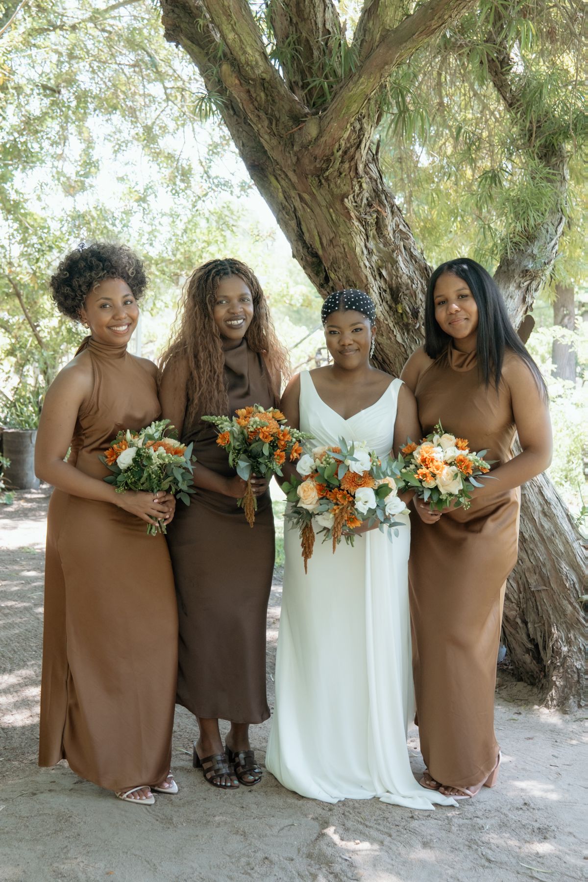 Bride in white dress poses with three bridesmaids in brown satin gowns holding orange and white bouquets outdoors.