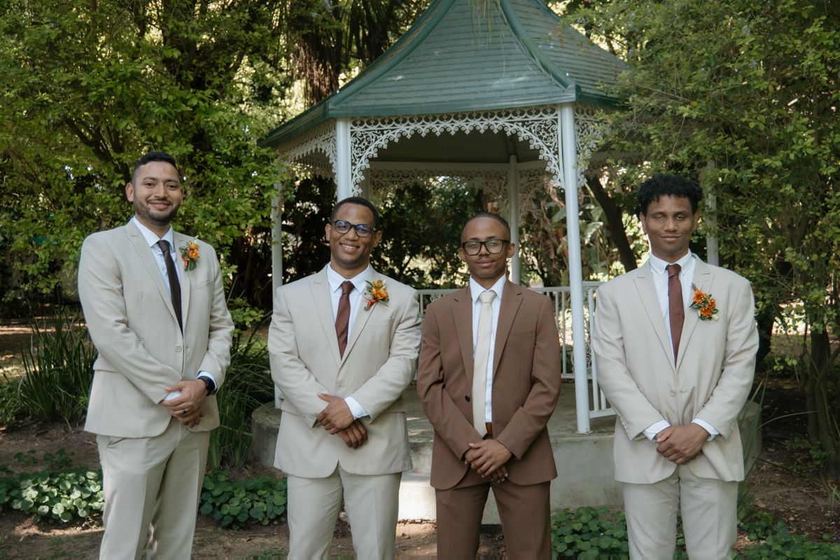 Four groomsmen in tan and brown suits pose together in front of white garden gazebo.