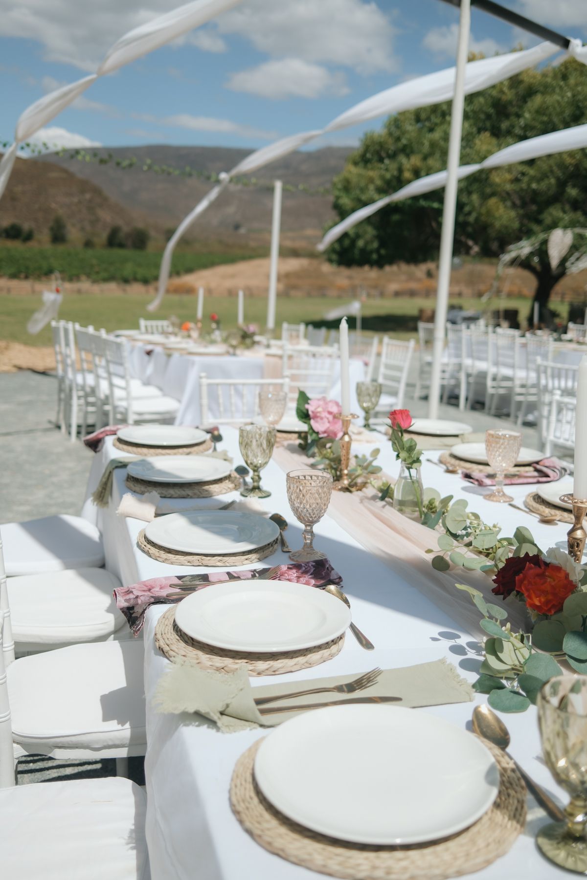 Long outdoor wedding reception table with white linens, wicker chargers, pink roses, and mountain backdrop.