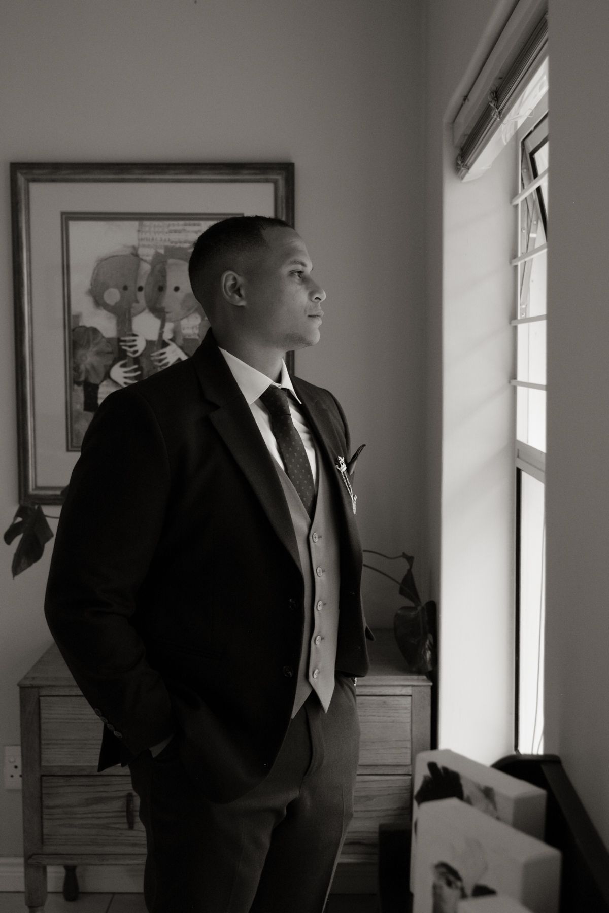 Dapper groom in dark suit gazing thoughtfully out a window in black and white portrait.
