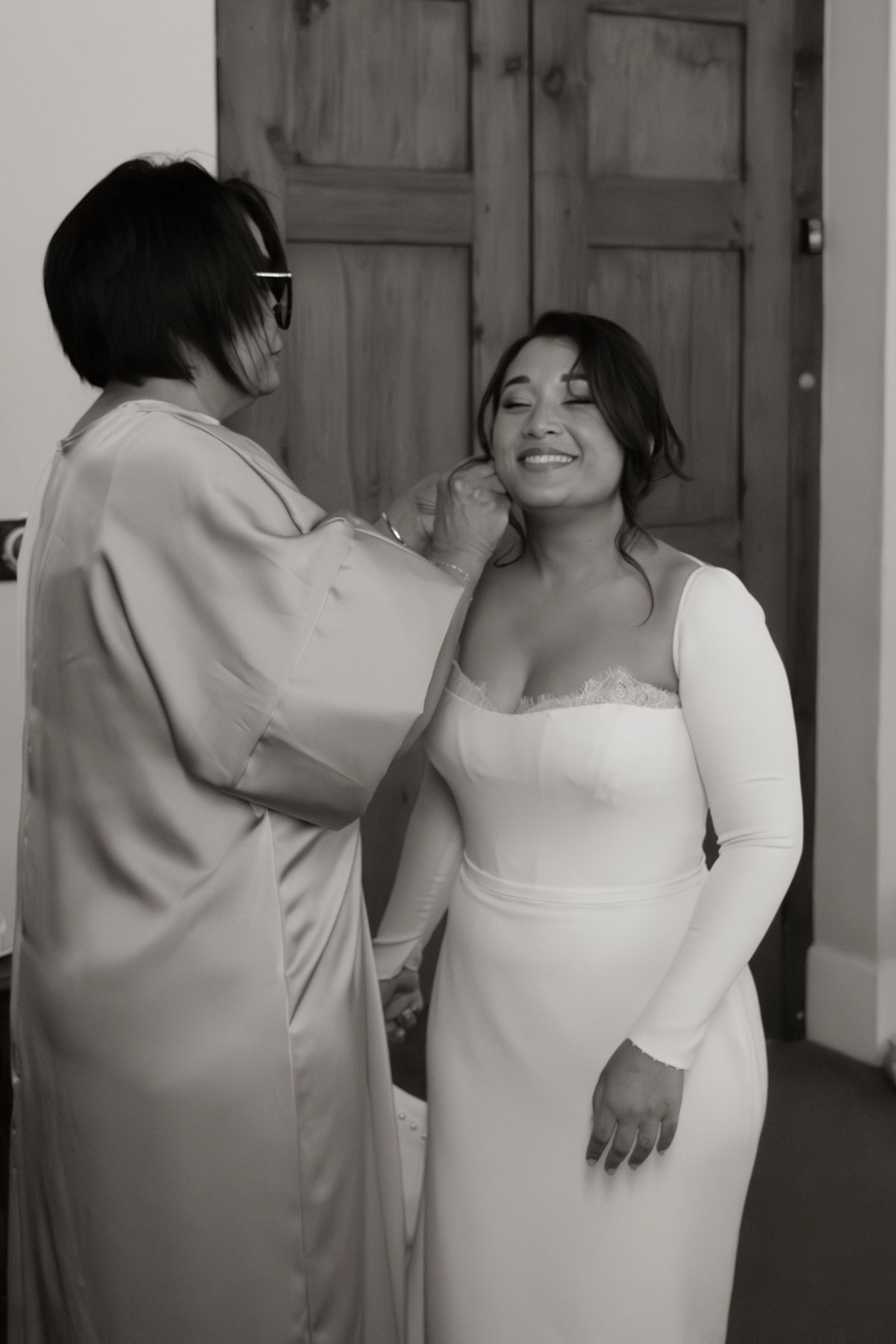 Black and white photo of a woman helping a smiling bride in a white gown with her jewelry before the wedding.