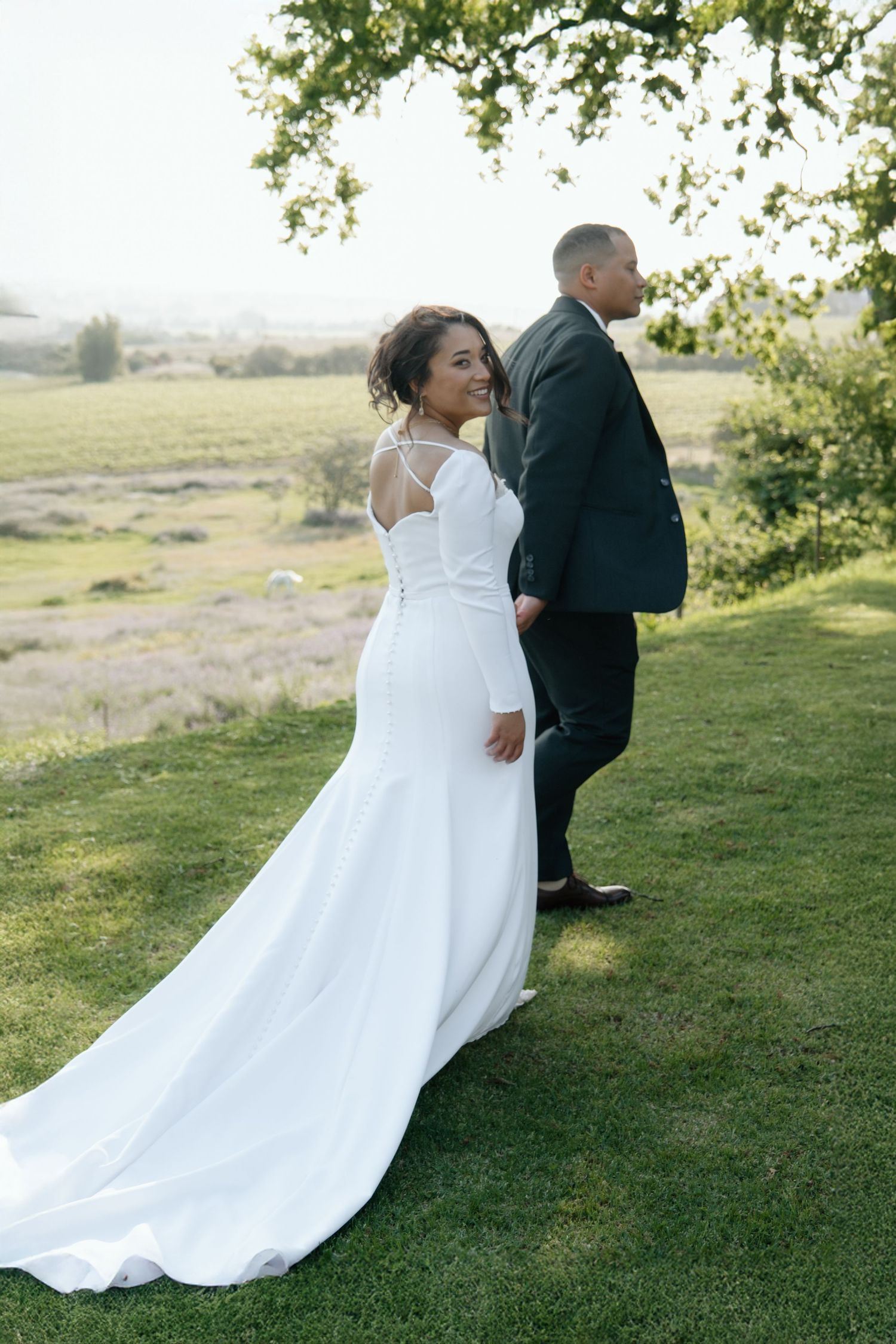Smiling bride in elegant white gown with train holds grooms hand in lush green outdoor setting.