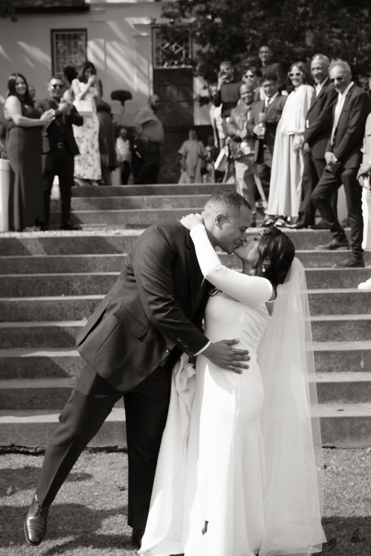 Groom dips bride for romantic kiss on church steps surrounded by wedding guests in black and white.
