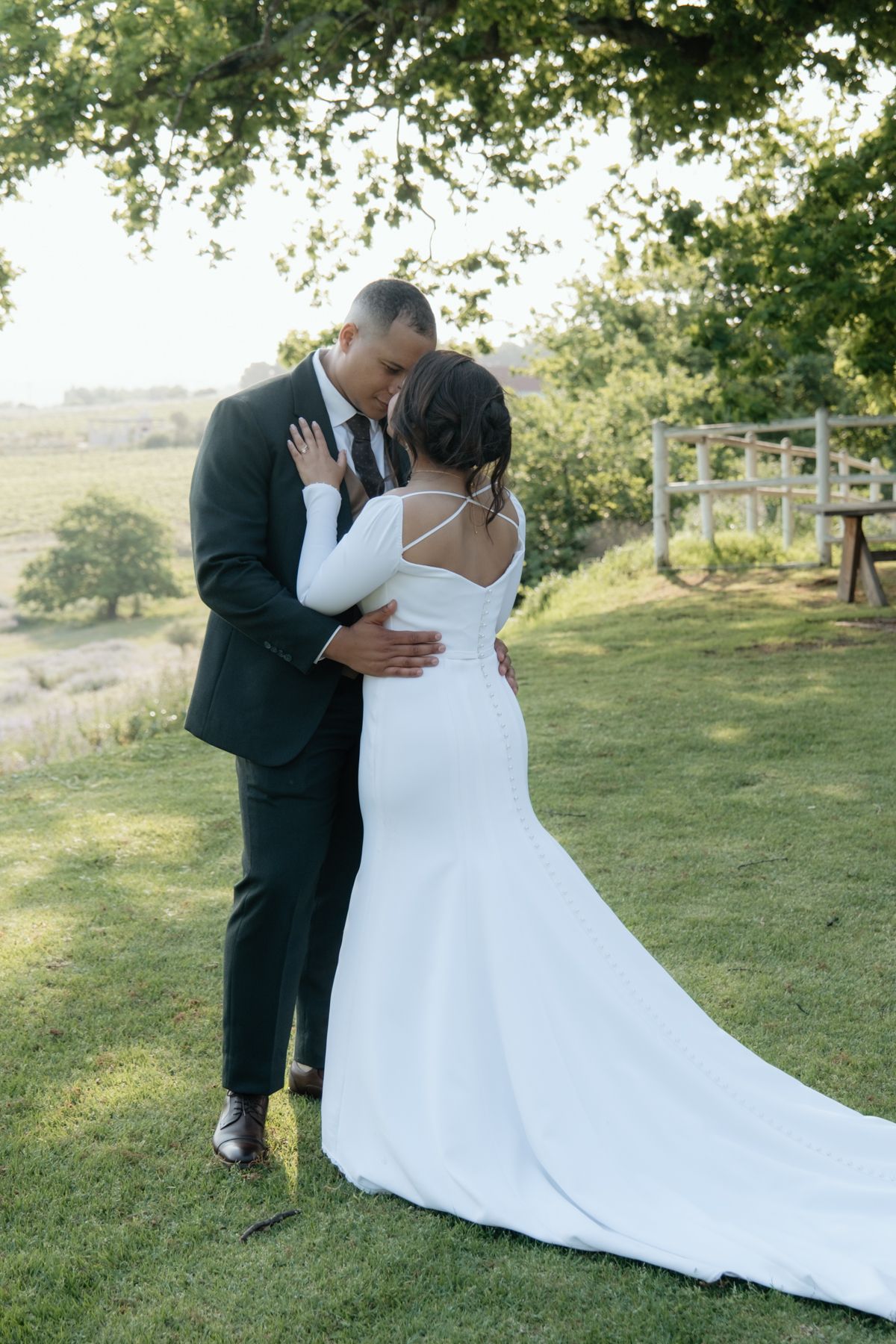 Bride and groom sharing romantic embrace under oak tree on green lawn with countryside views at golden hour.