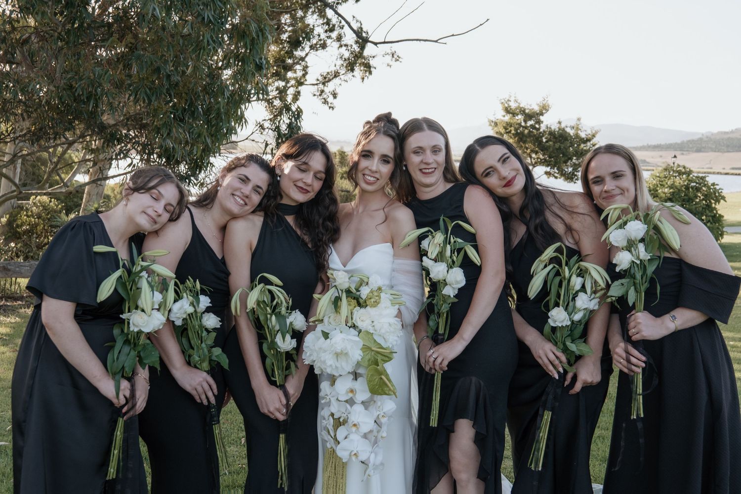 Wedding party in black dresses posing together with white floral bouquets outdoors.
