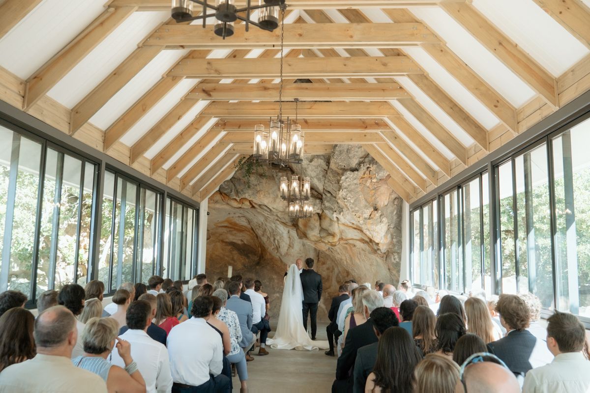 Elegant wedding ceremony in a rustic chapel with wood-beam ceilings, chandelier, and stunning natural rock wall backdrop.