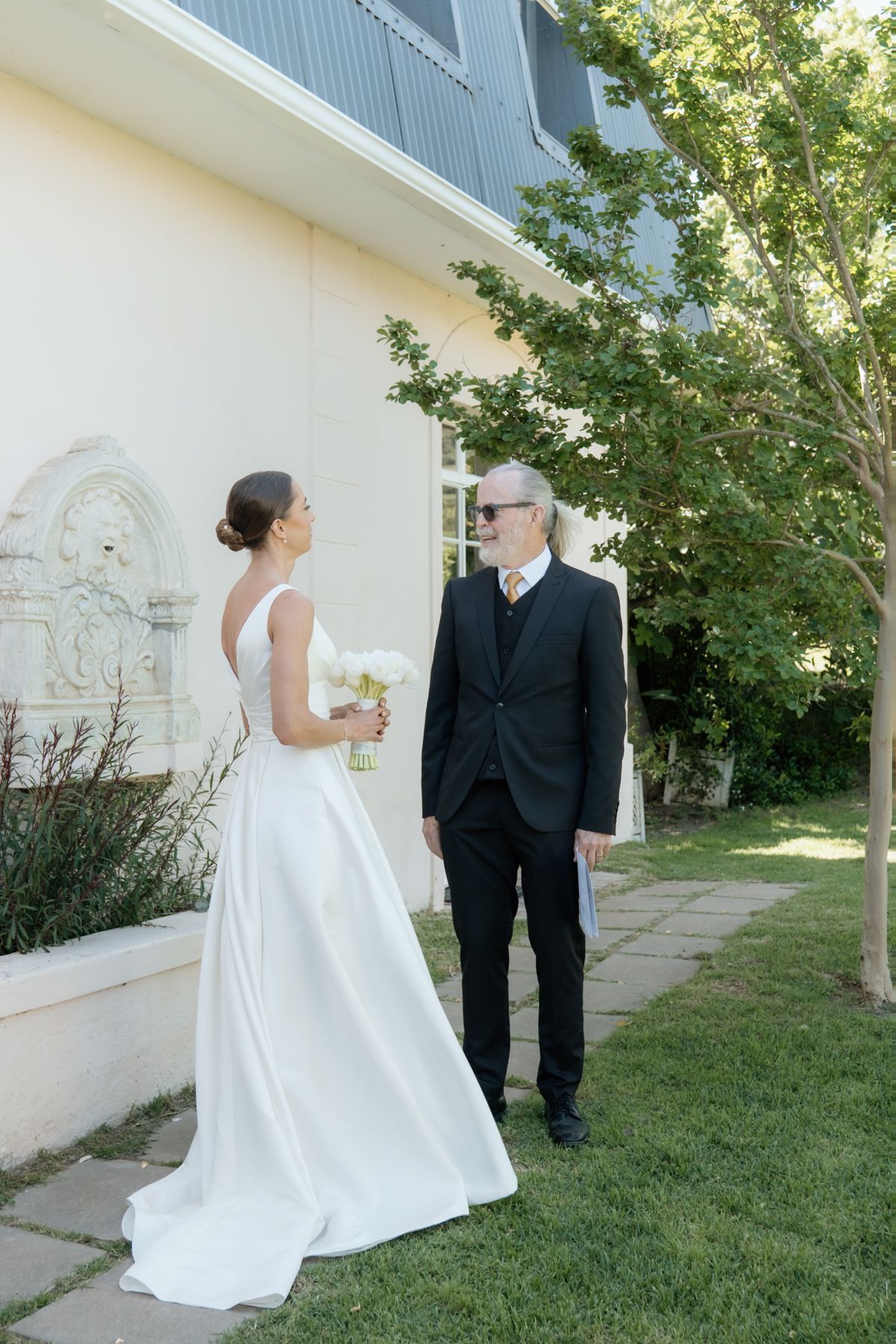 Bride in a white gown holds a bouquet while walking with an older gentleman in black suit outdoors.