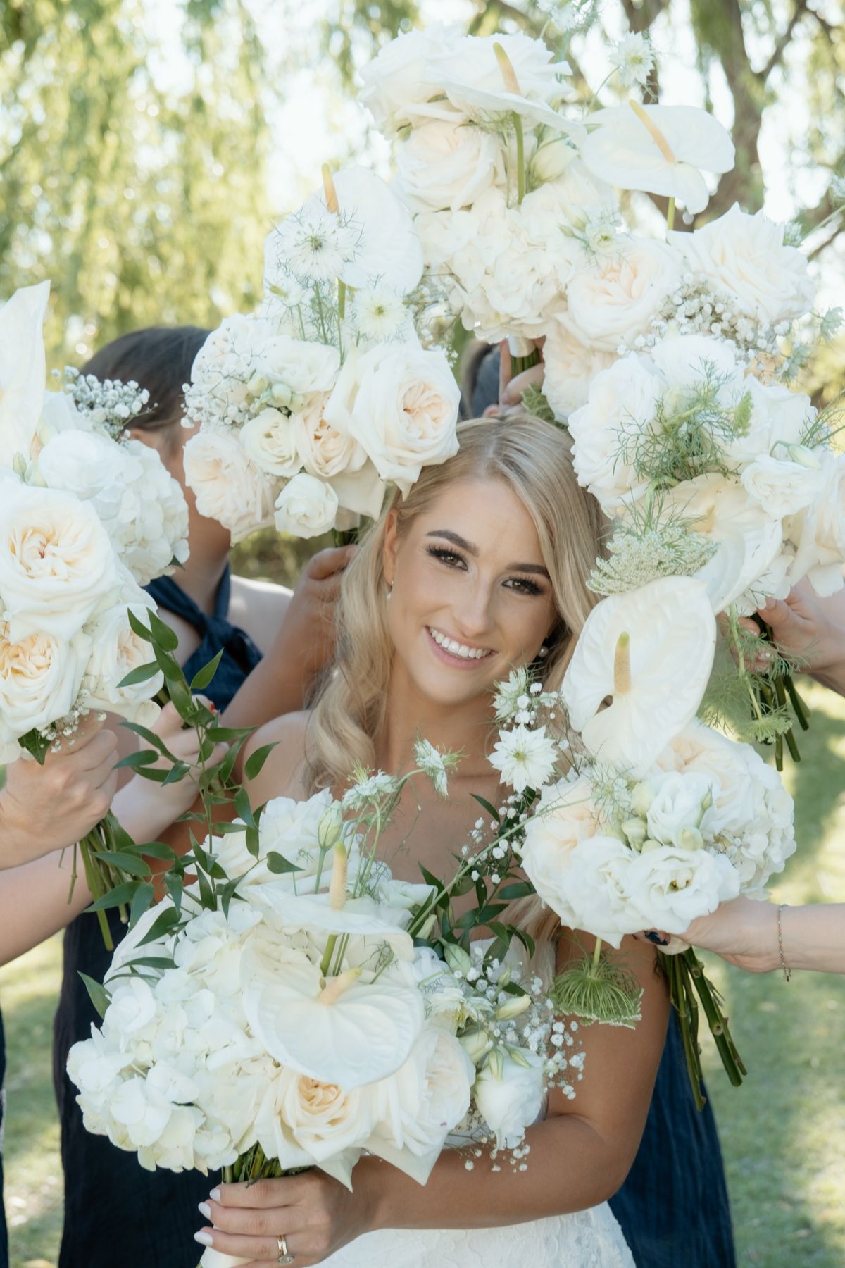 Smiling blonde bride surrounded by white floral bouquets of roses and dahlias held by bridesmaids outdoors.