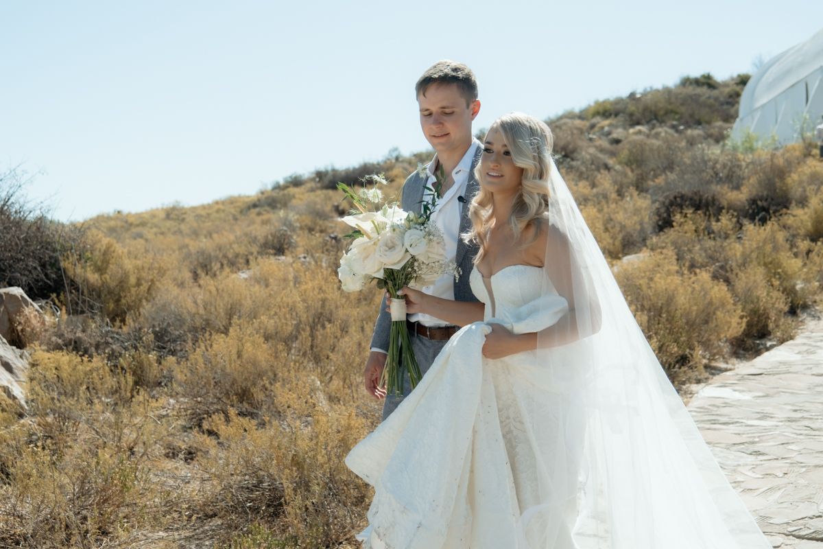 Bride and groom pose outdoors in desert landscape, bride holding white floral bouquet in strapless gown.