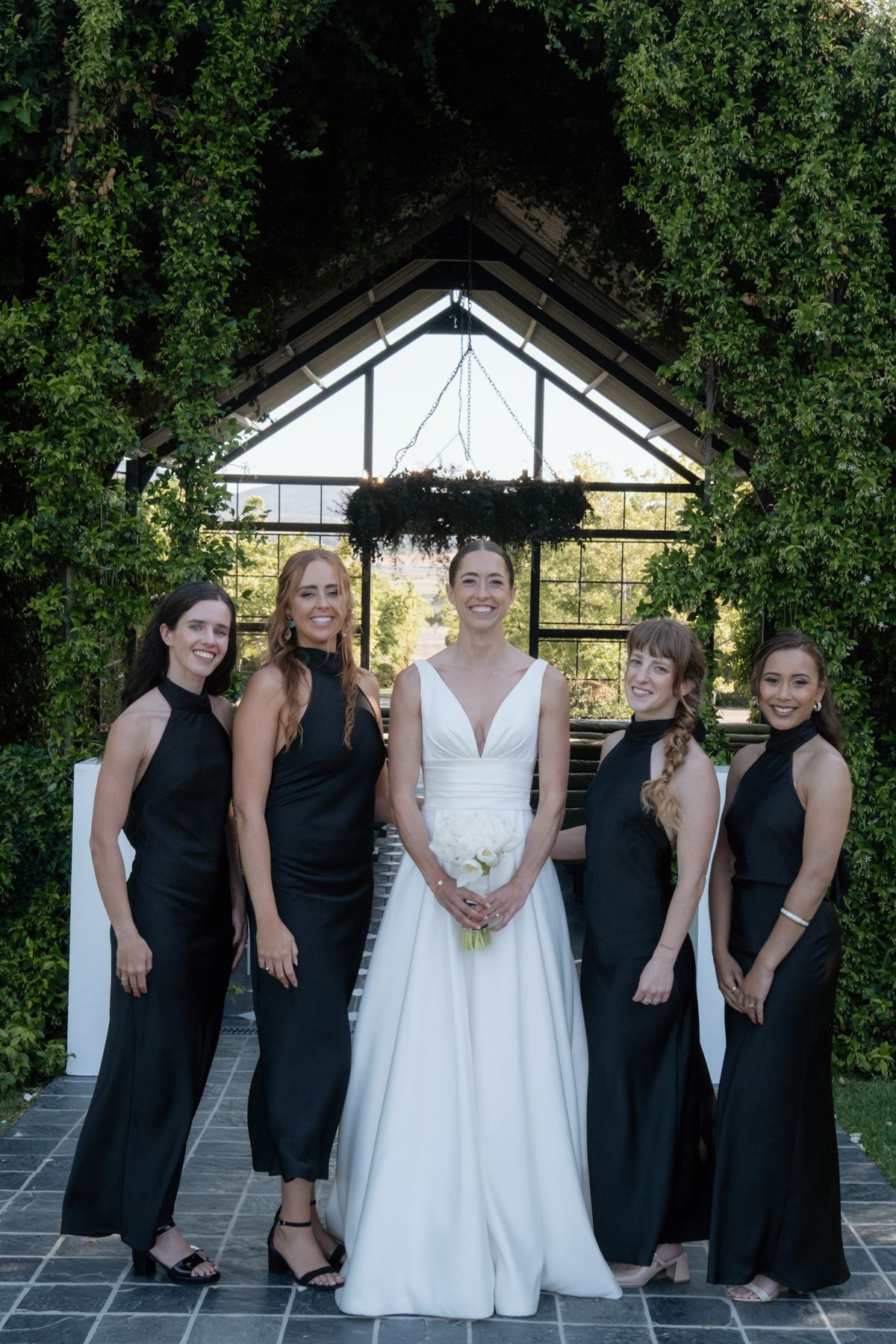 Bride in white V-neck gown poses with four bridesmaids in black dresses before a lush green archway backdrop.