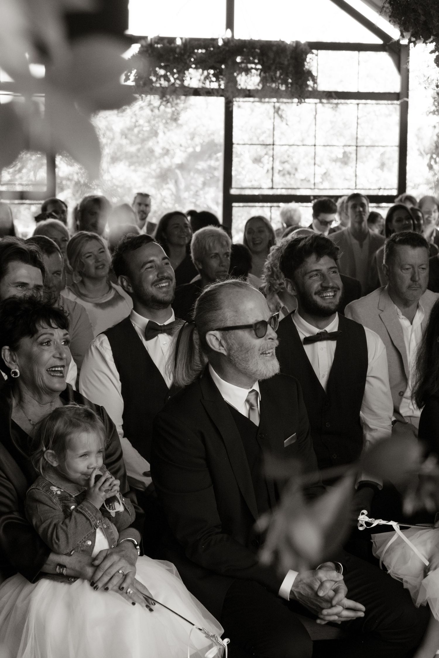 Black and white photo of smiling wedding guests seated during an indoor ceremony.